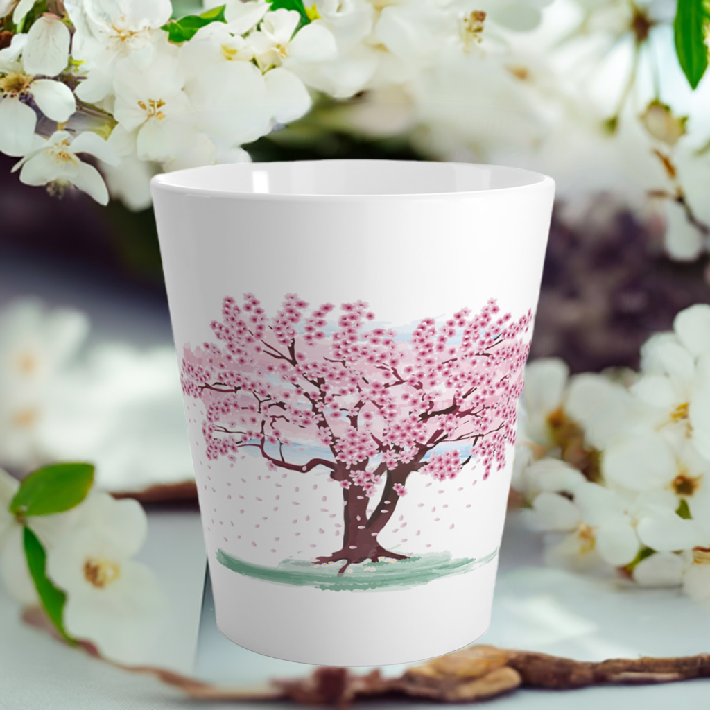 A white latte mug with a pink blossoming tree design, placed on a white ceramic surface with white flowers in the background.