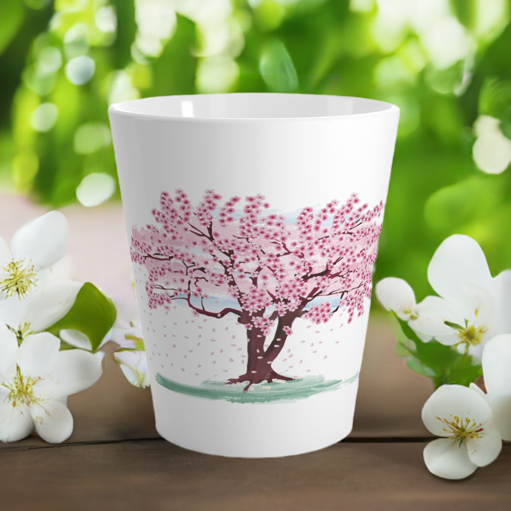A white latte mug with a pink blossoming tree design, placed on a wooden surface with white and green flowers in the background.