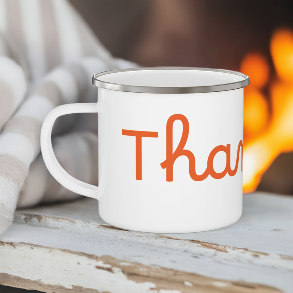 A white stainless steel camping mug with orange 'THANKFUL' text placed on a white wooden surface with a campfire and blanket background.